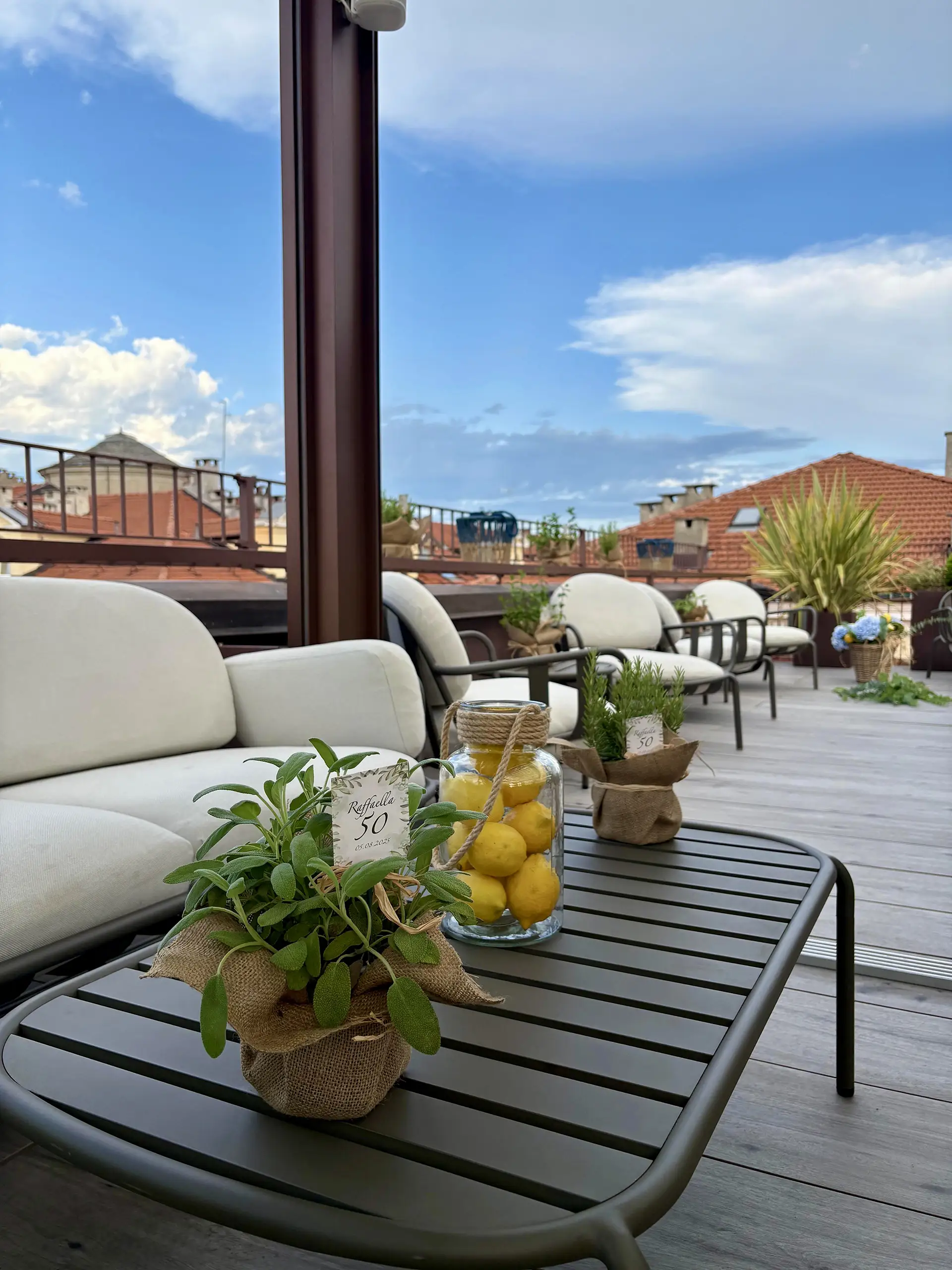Nell'immagine c'è la terrazza panoramica dell'hotel. In primo piano c'è un tavolino nero basso con sopra un vasetto di salvia, un vasetto trasparente con dentro dei limoni e un vasetto di rosmarino. A sinistra c'è un divanetto bianco. Dietro ci sono delle sedie bianche morbide e in fondo, nell'angolo, un vaso di con una pianta verde. Oltre il parapetto della terrazza ci sono i tetti delle case accanto.