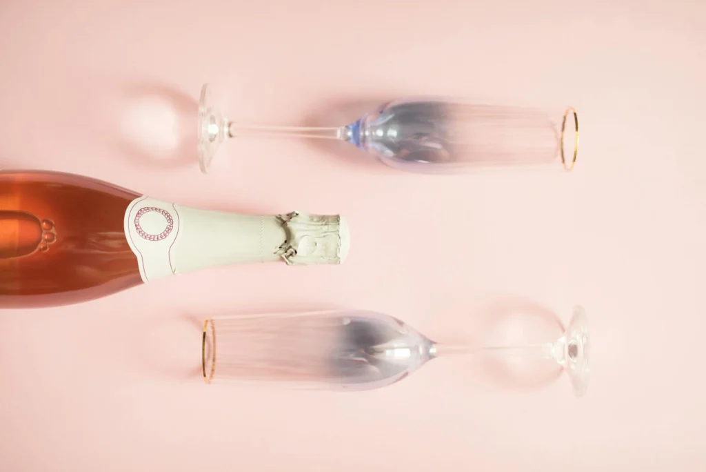 Nell'immagine a sinistra al centro c'è una bottiglia di champagne rosè sdraiata su un piano rosa e su entrambi i lati della bottiglia due calici da spumante.