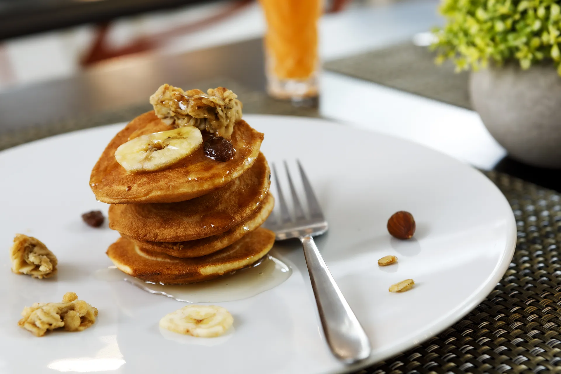 Dettaglio colazione hotel. Primo piano di piatto bianco con tre pancake impilati, guarniti con fettine di banana, granola e sciroppo. Accanto, forchetta in acciaio e qualche nocciola sparsa sul piatto. Sullo sfondo sfocato, bicchiere con succo arancione e vaso con pianta decorativa.